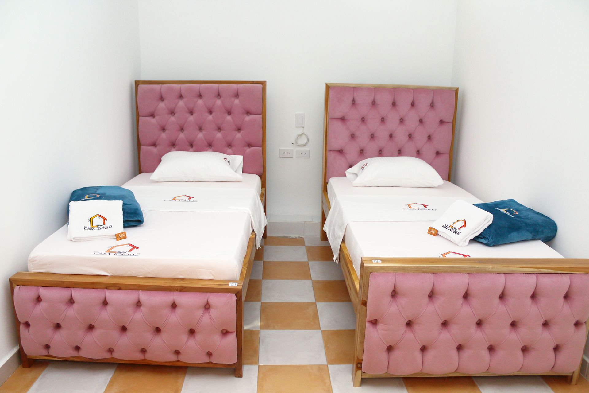 Dos camas individuales con cabeceros capitoné rosas en una habitación con paredes blancas y suelo de baldosas ajedrezadas.