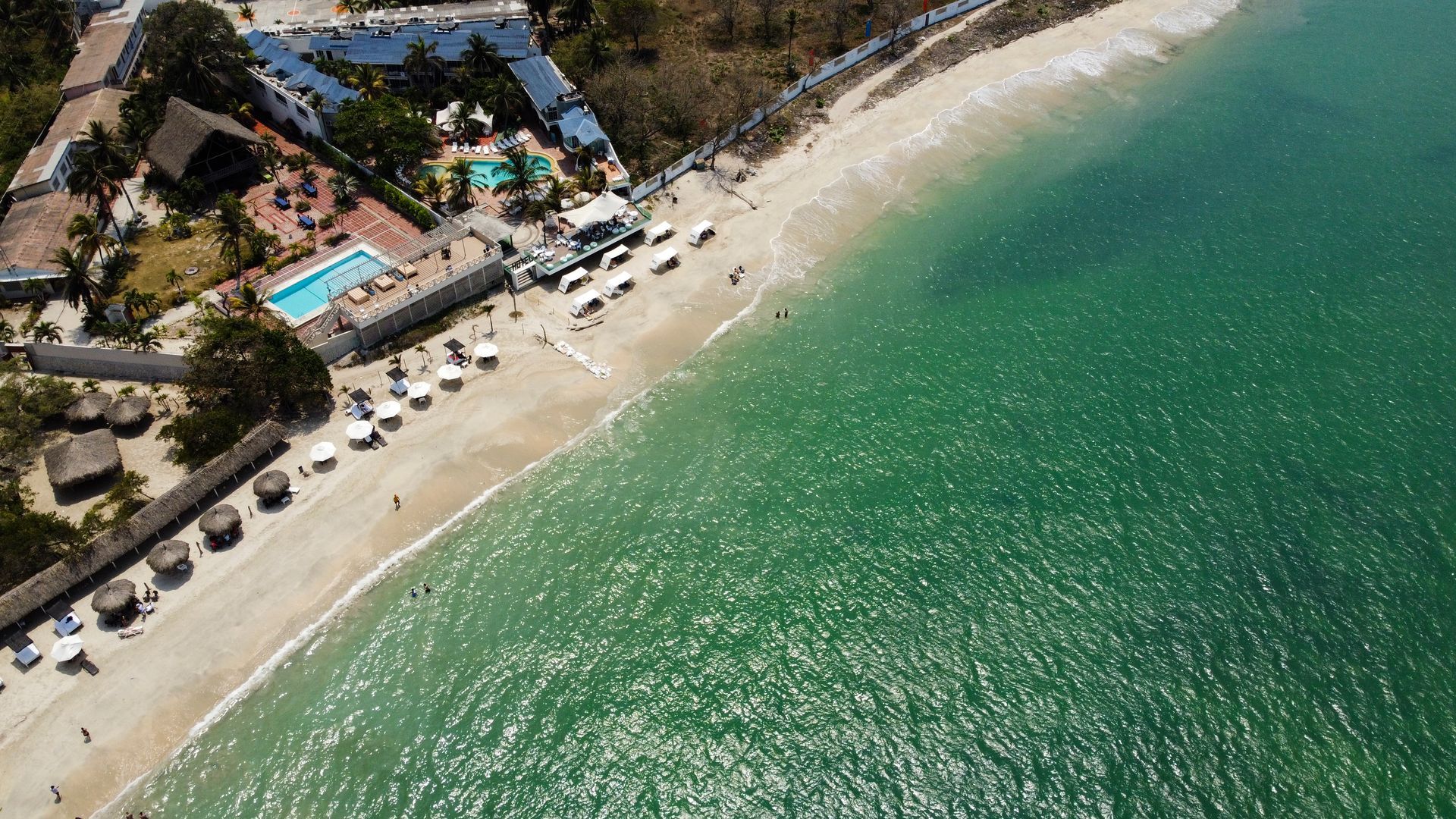 Vista aérea de la playa con el complejo turístico, agua turquesa y arena blanca.