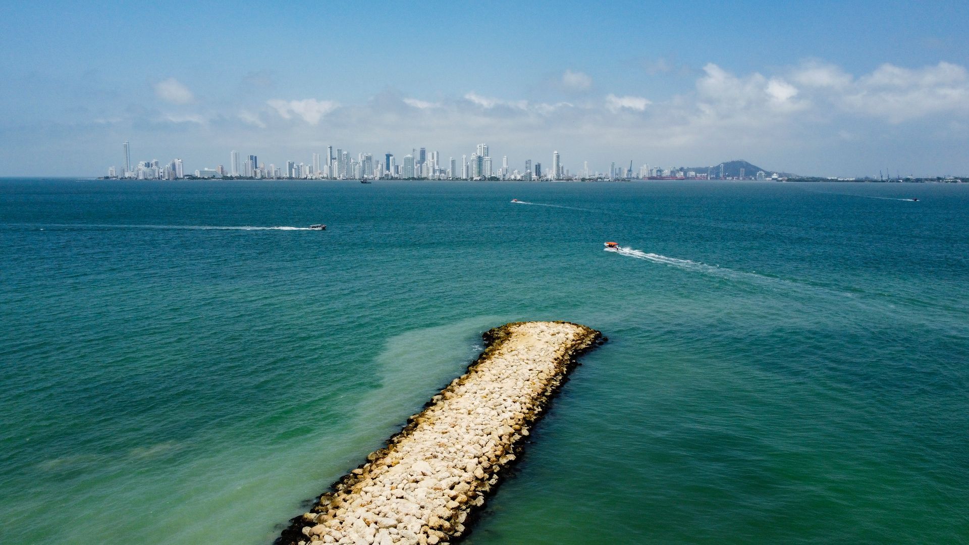 Vista al océano con el horizonte de la ciudad a lo lejos; un muelle rocoso se adentra en las aguas turquesas.