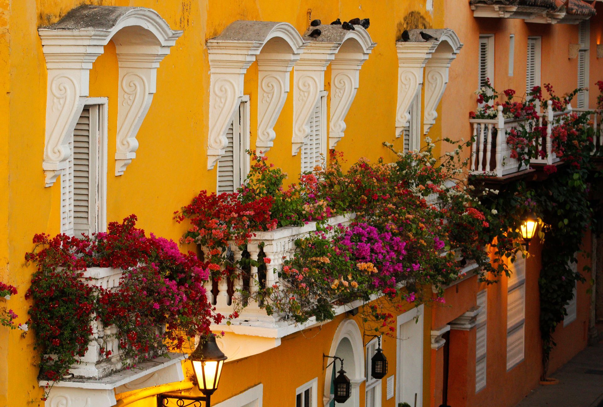 Fachadas de edificios amarillas y naranjas con balcones rebosantes de flores coloridas, estilo colonial.