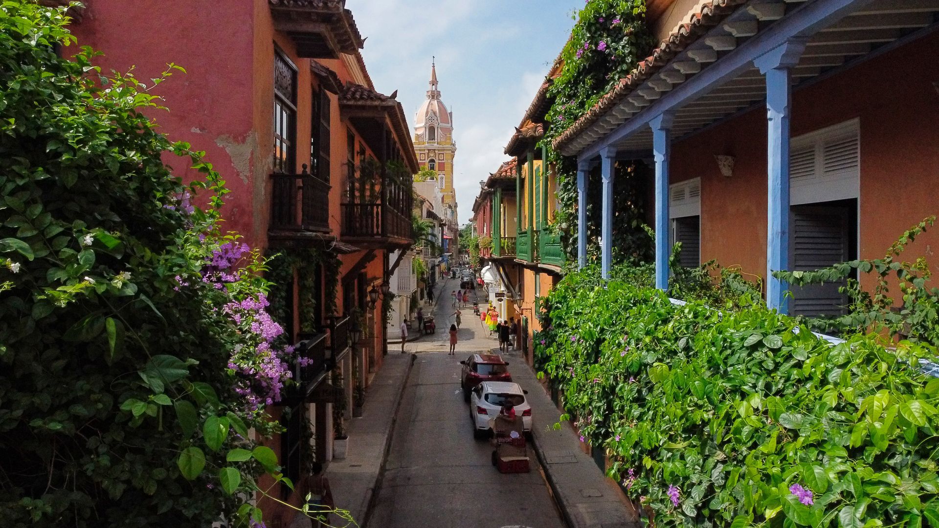 Calle estrecha de Cartagena de Indias, Colombia, bordeada de coloridos edificios coloniales, vegetación y coches.