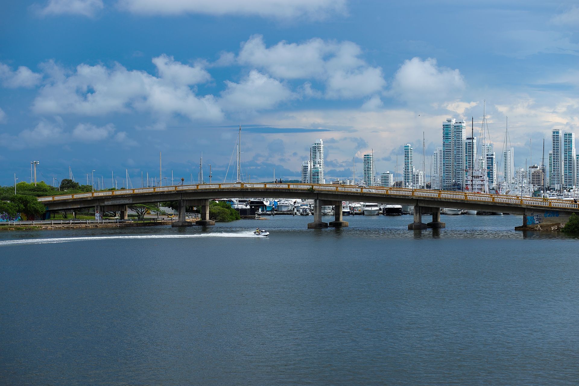 Puente sobre el agua con el horizonte de la ciudad y barcos al fondo bajo un cielo nublado.