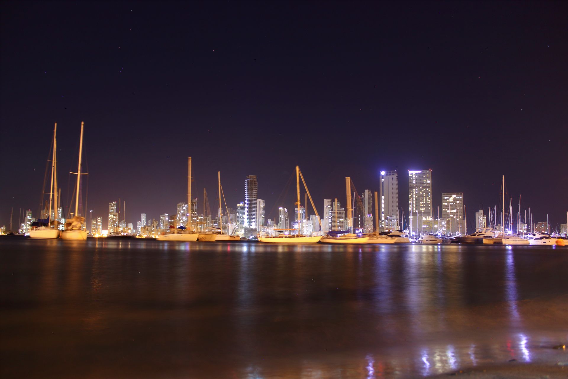 Paisaje urbano nocturno con edificios iluminados y veleros en una bahía tranquila.