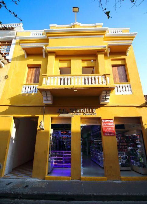 Edificio amarillo con escaparates; balcón con barandilla blanca, cielo azul.