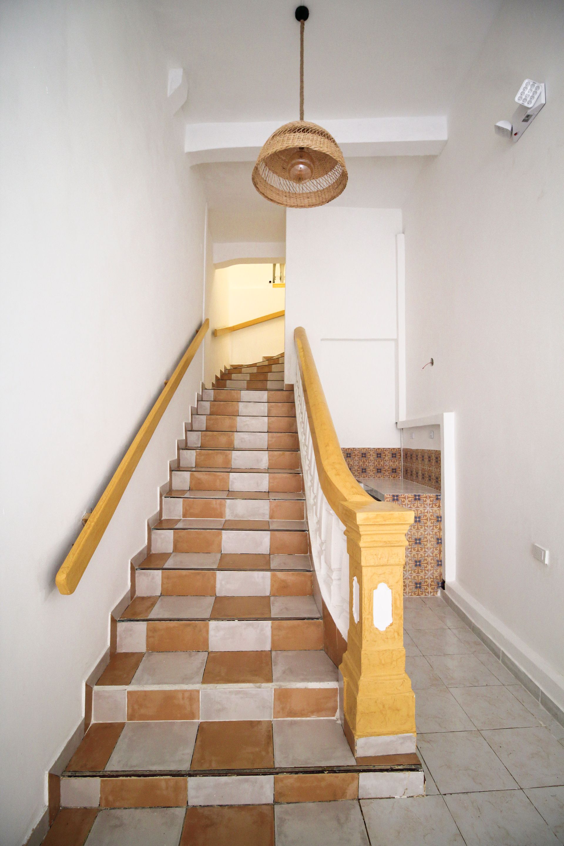 Escalera con peldaños de baldosas ajedrezadas, pasamanos de madera y lámpara colgante de mimbre.
