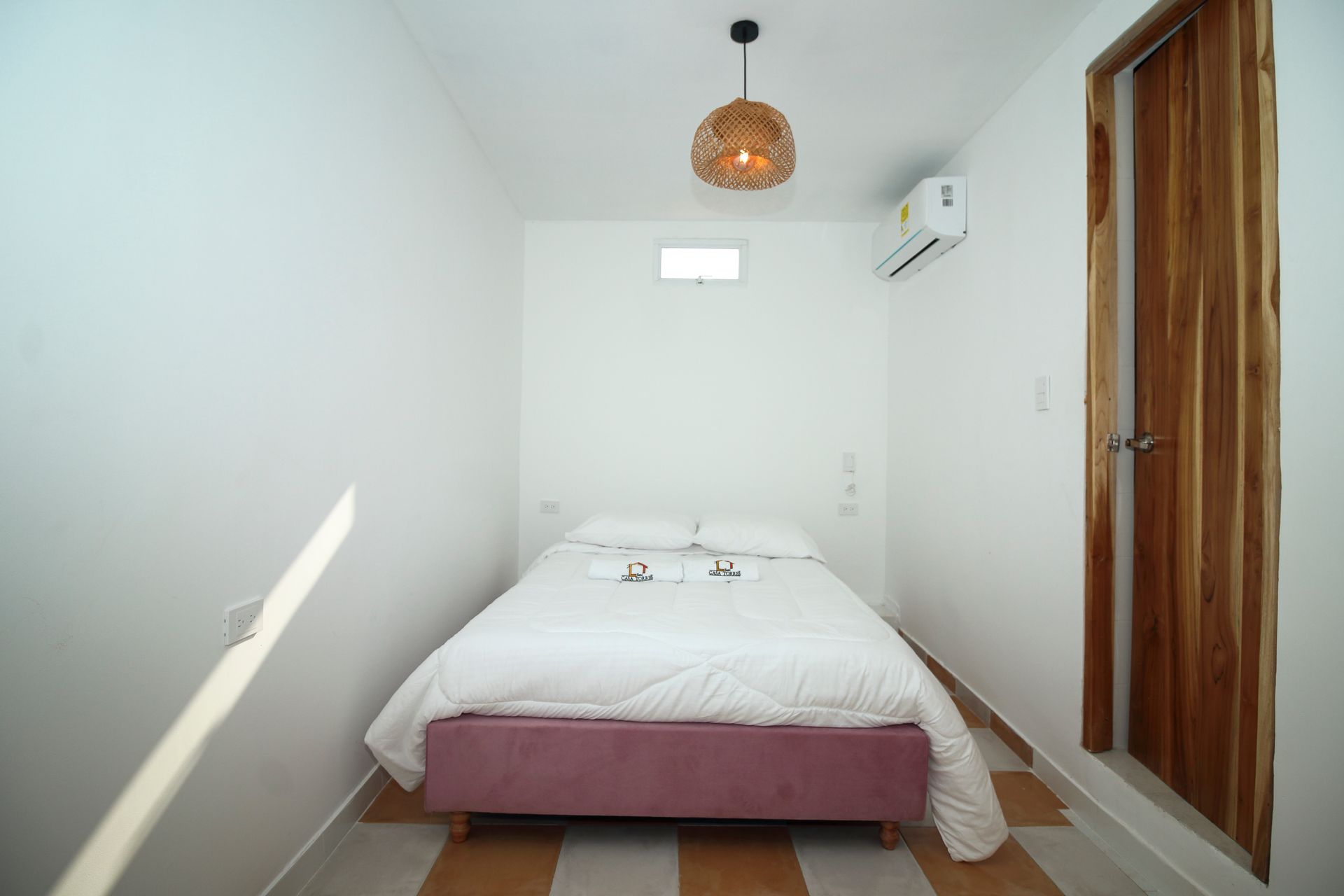 Small, white bedroom with bed, wooden door, and air conditioner; woven light fixture.