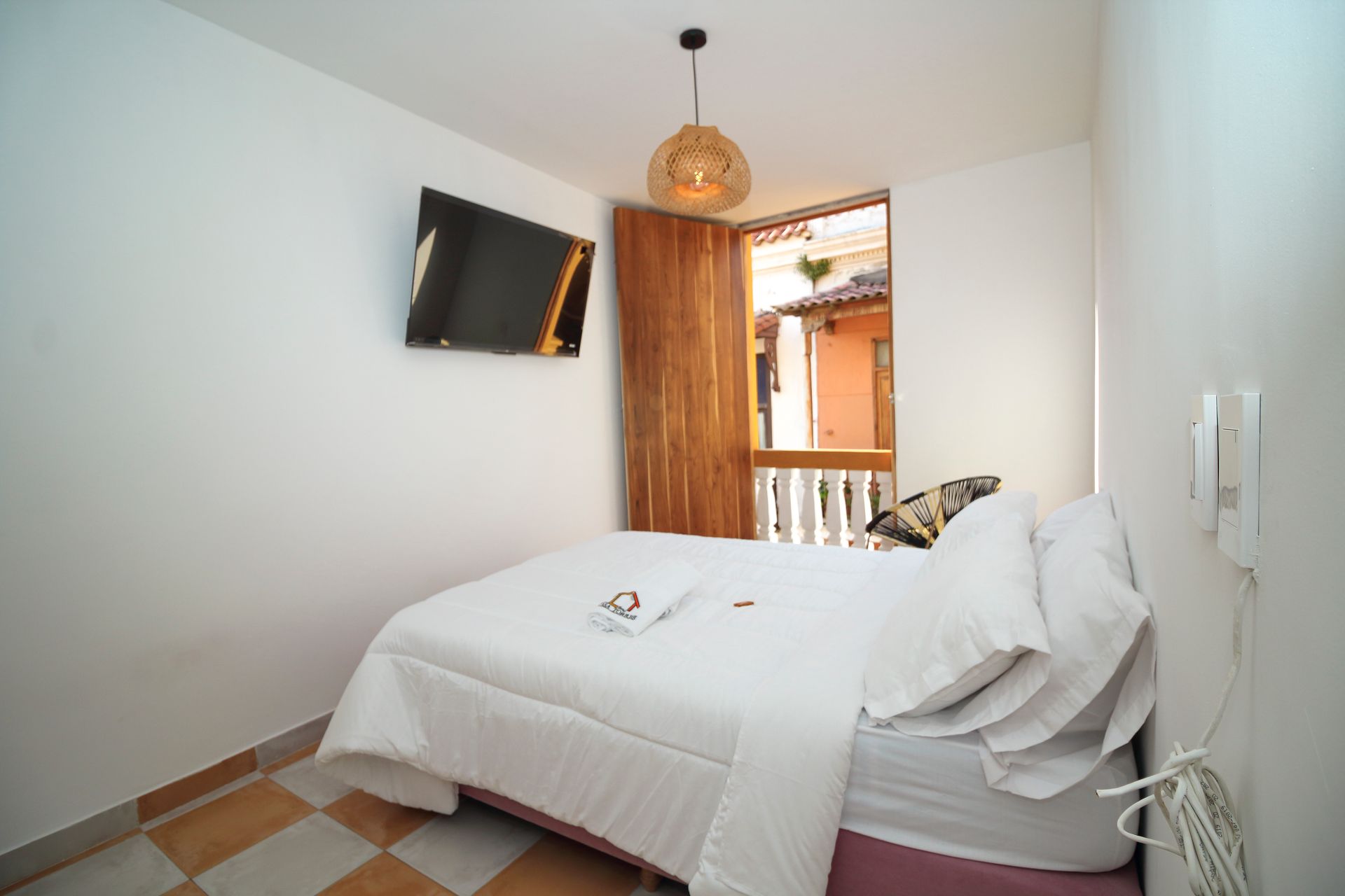 Small hotel room with bed, TV, and open window looking out to a balcony and other buildings.