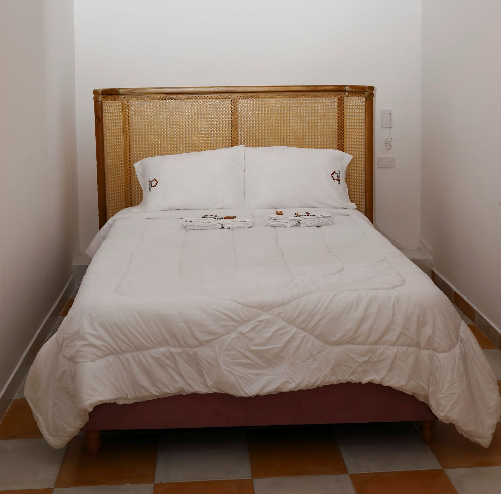 Dormitorio con cama, ropa de cama blanca, cabecero de mimbre y suelo ajedrezado.