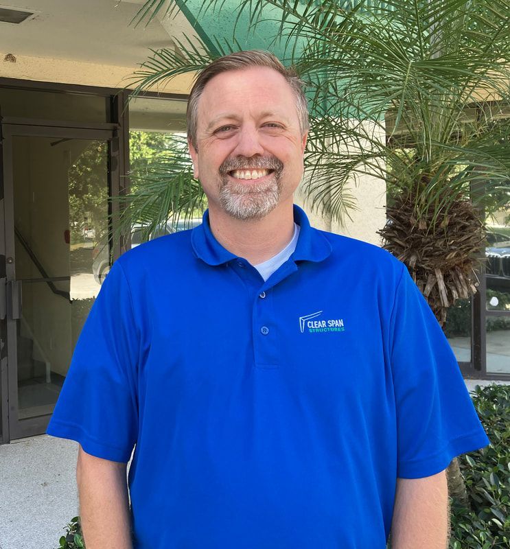 A man with a beard wearing a blue shirt is smiling in front of a building.