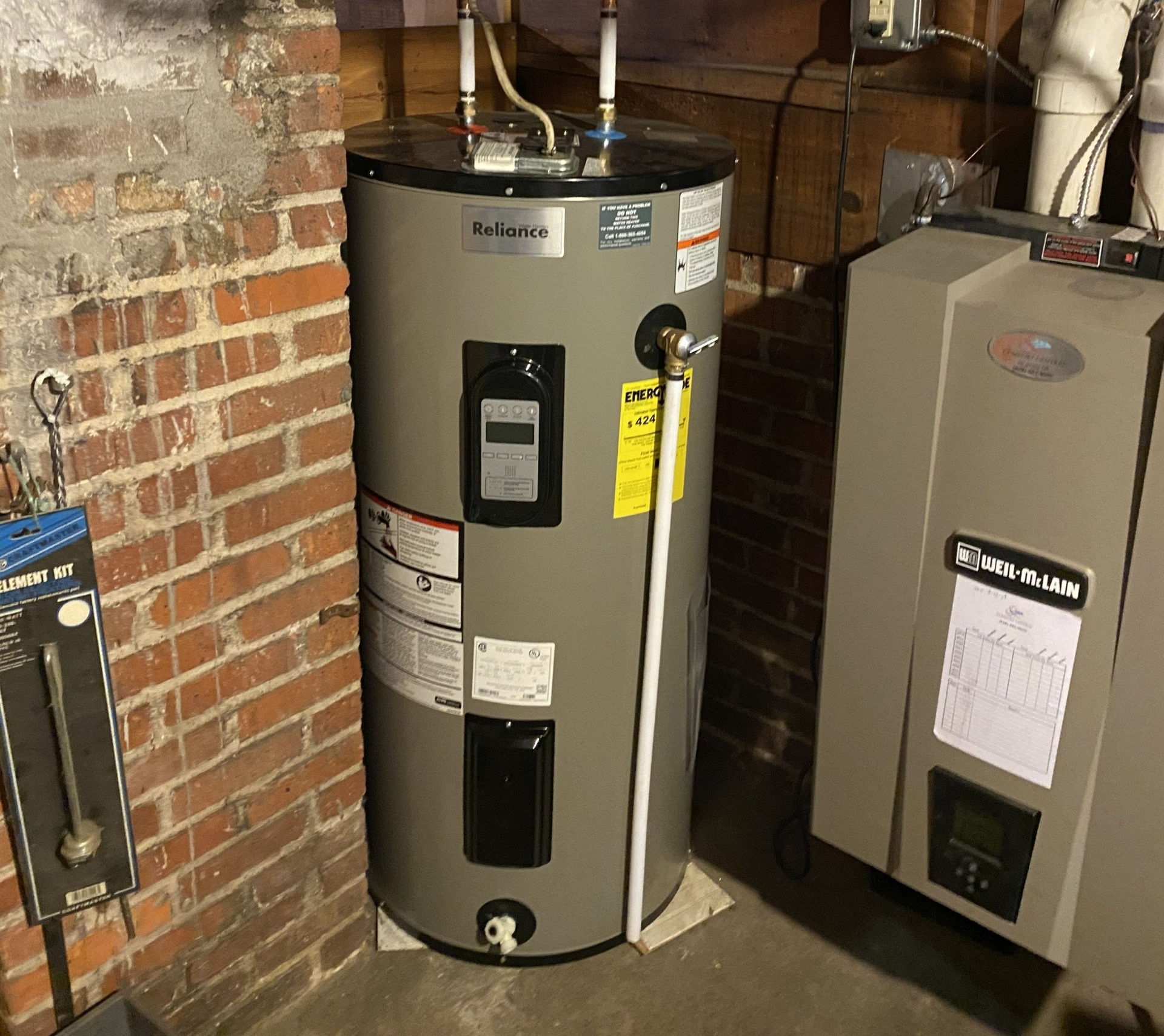 A water heater is sitting in a basement next to a brick wall.