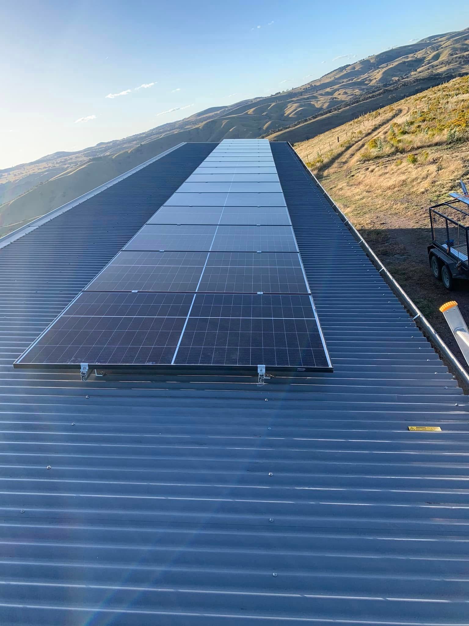 Solar panels on a corrugated metal roof beside a hillside under clear blue sky.