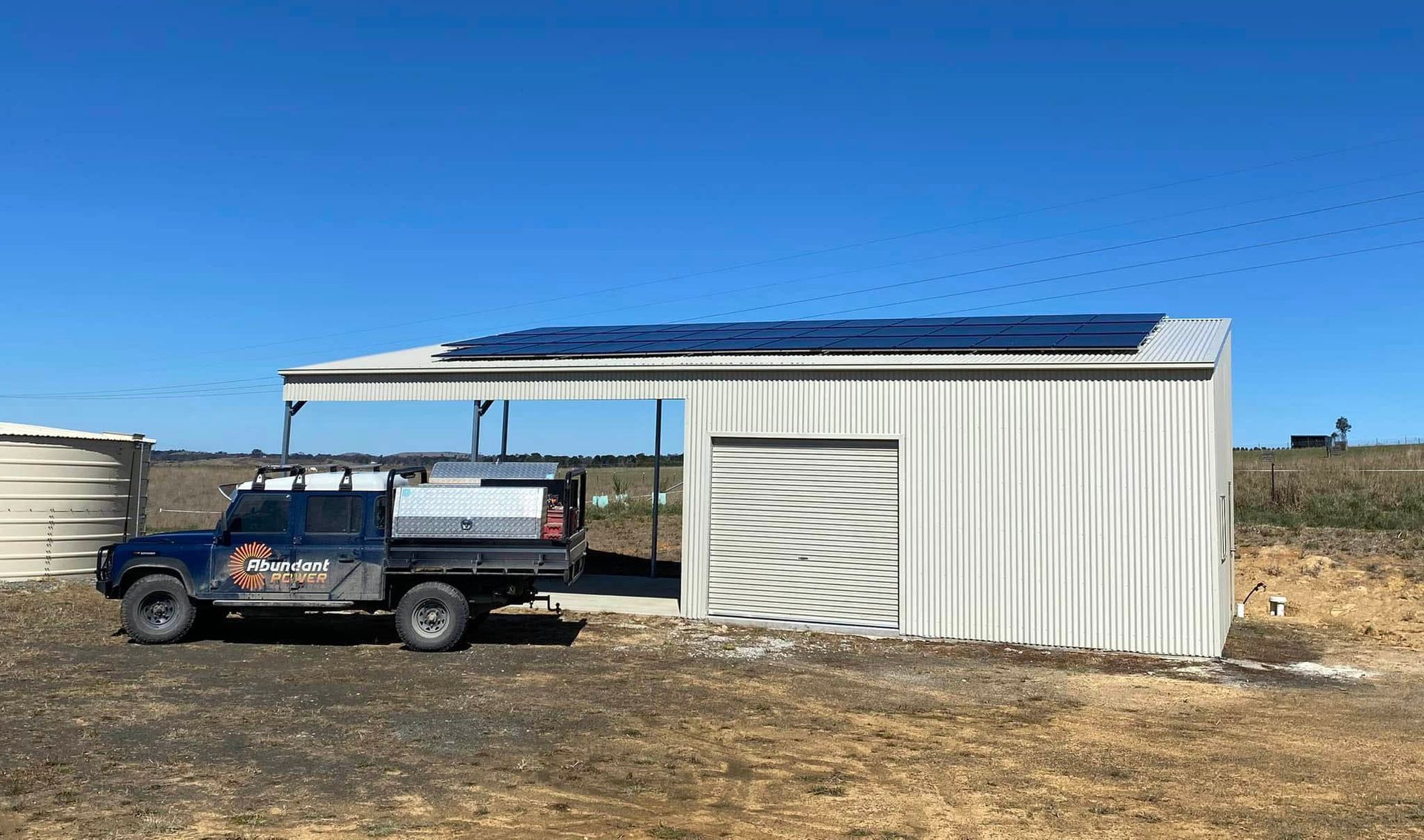 Blue pickup truck parked beside a white metal shed in a dry field under a clear sky