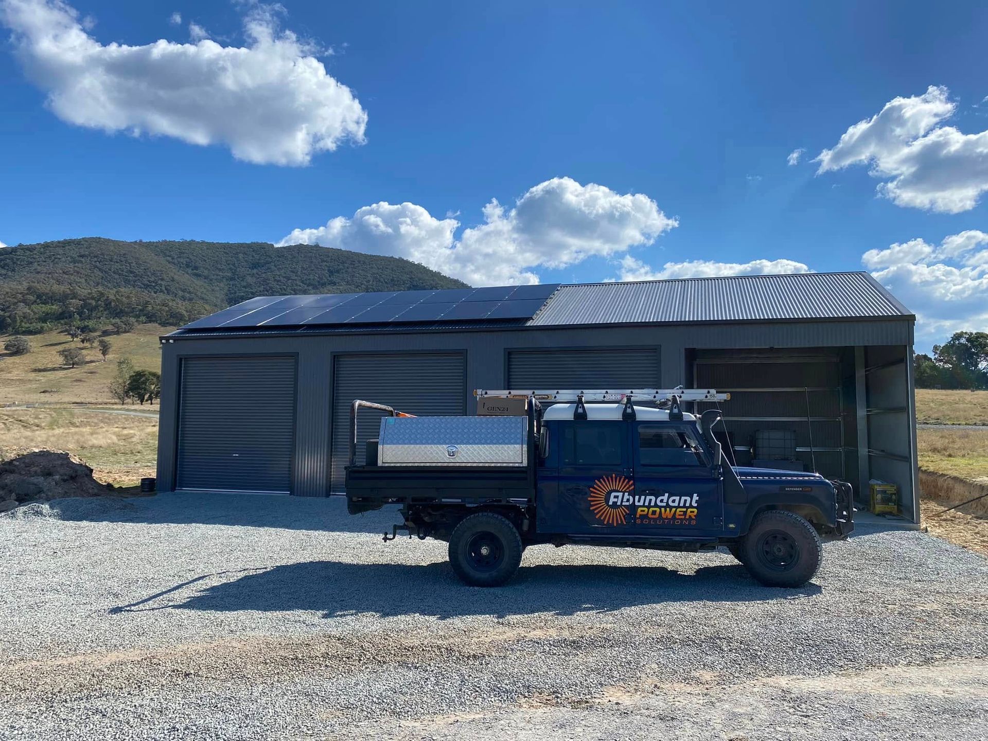 Blue utility truck parked beside a gray metal shed under a bright blue sky in a rural gravel lot