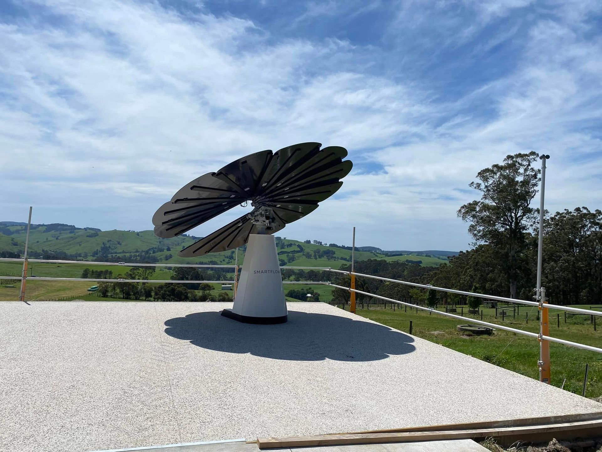 Solar-powered wind turbine sculpture on a gravel platform overlooking green fields and blue sky