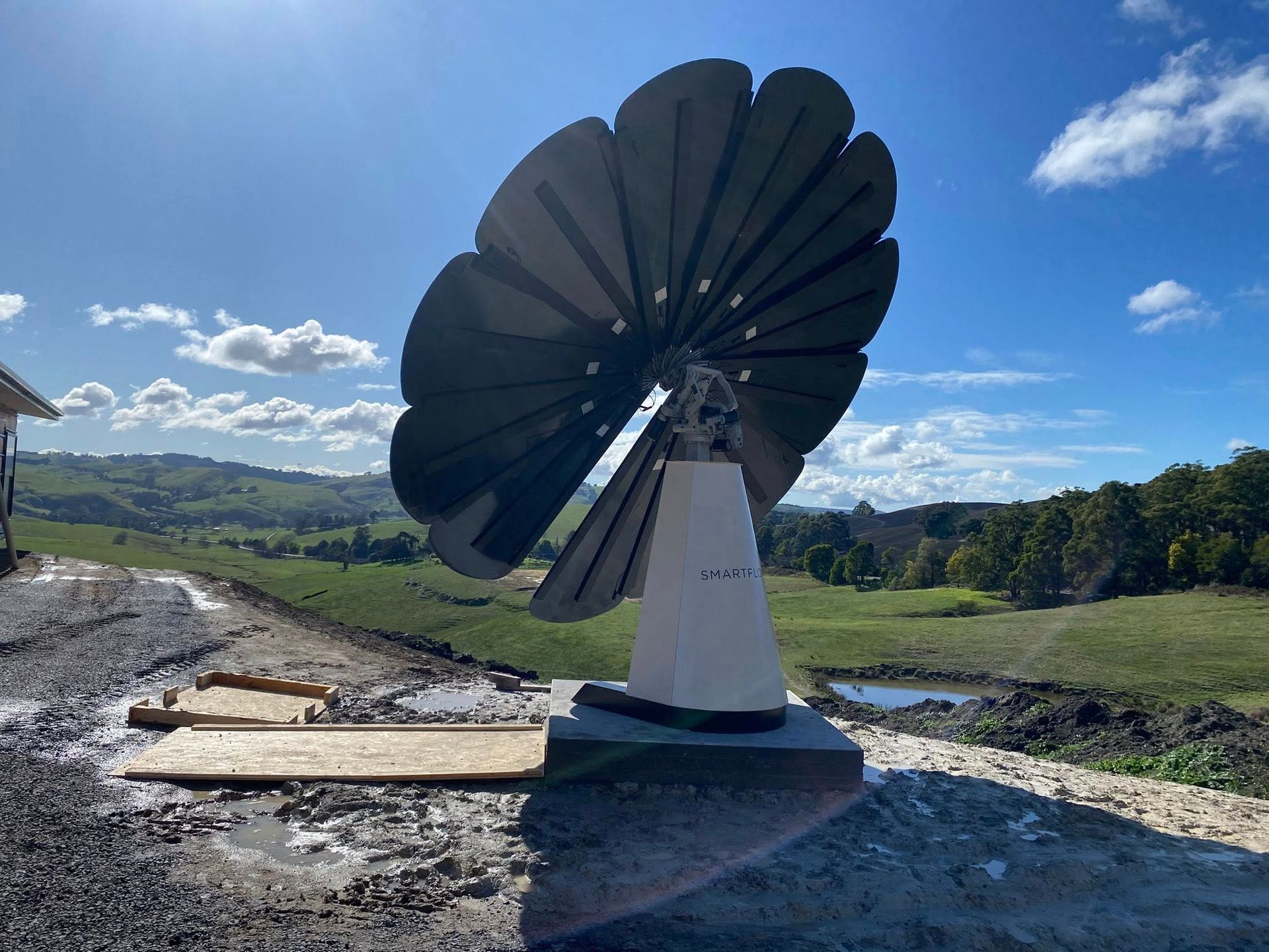 Large black wind turbine on a gravel site under a blue sky with clouds, overlooking green fields
