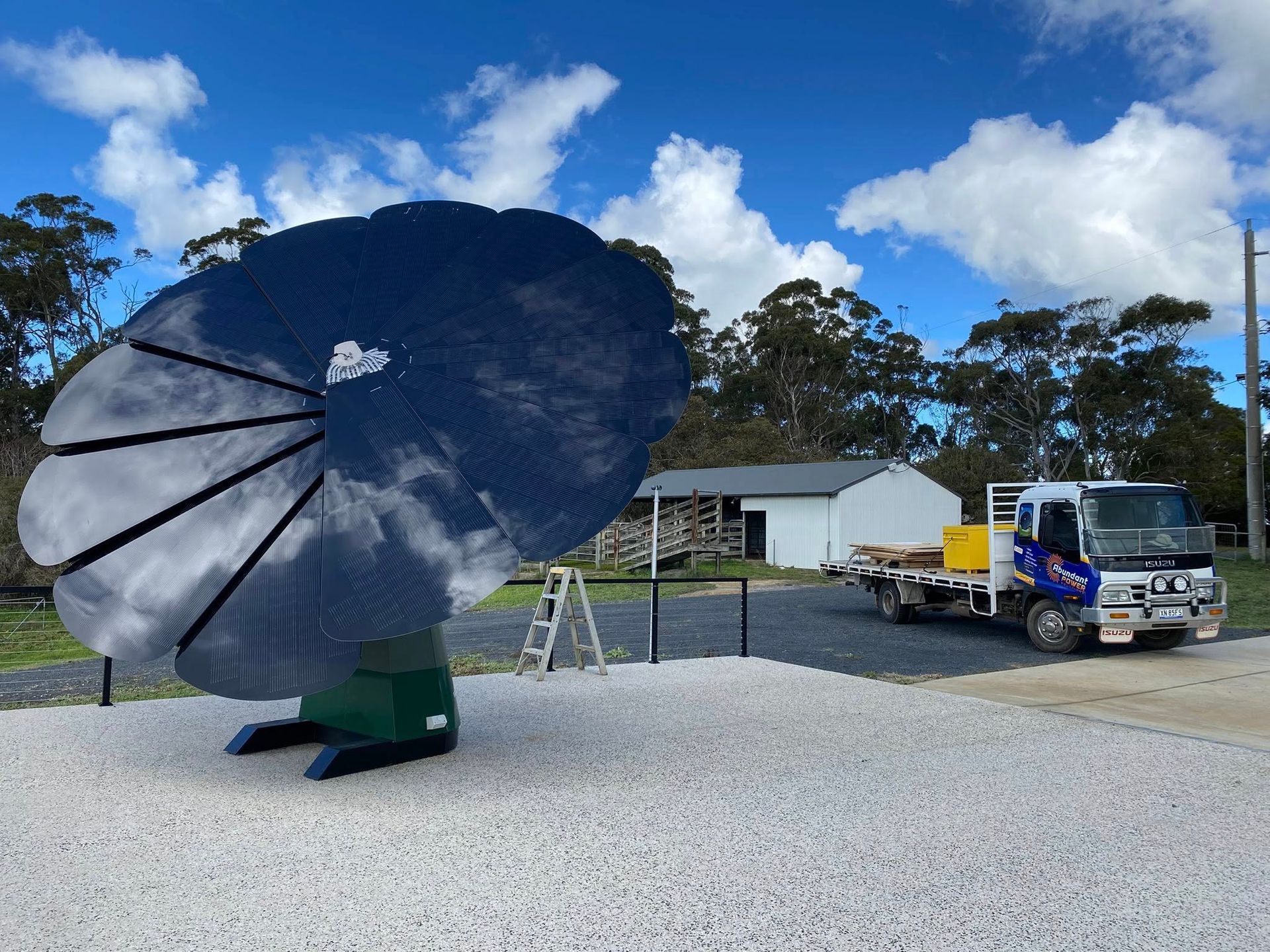 Large black satellite dish on a gravel lot with trucks, a ladder, and a small white building under a blue sky