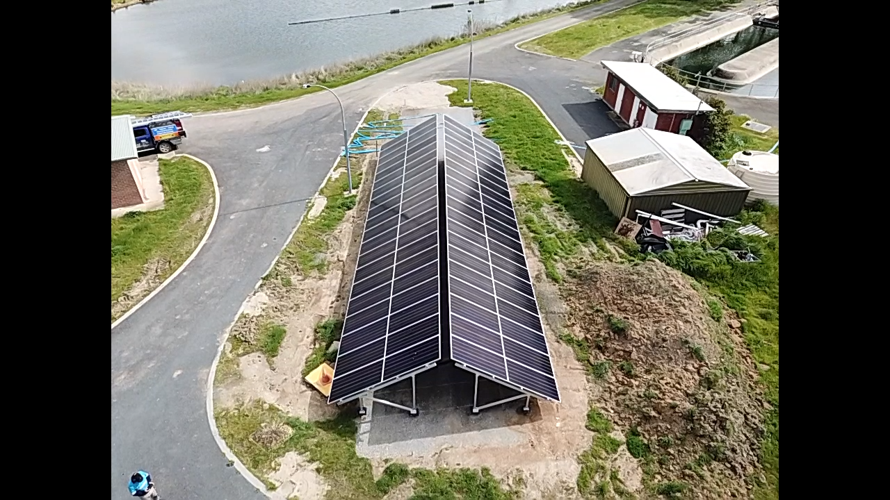 Aerial view of long solar panel arrays beside a curving road and water.