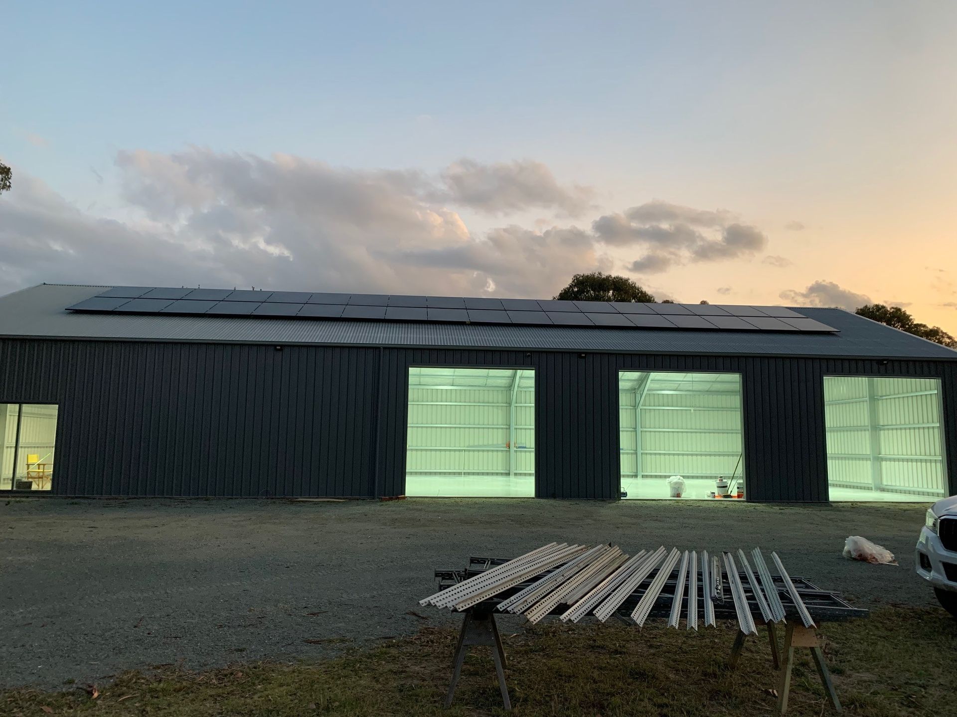 Large dark warehouse at dusk with lit garage doors, solar panels on roof, and stacked lumber in front