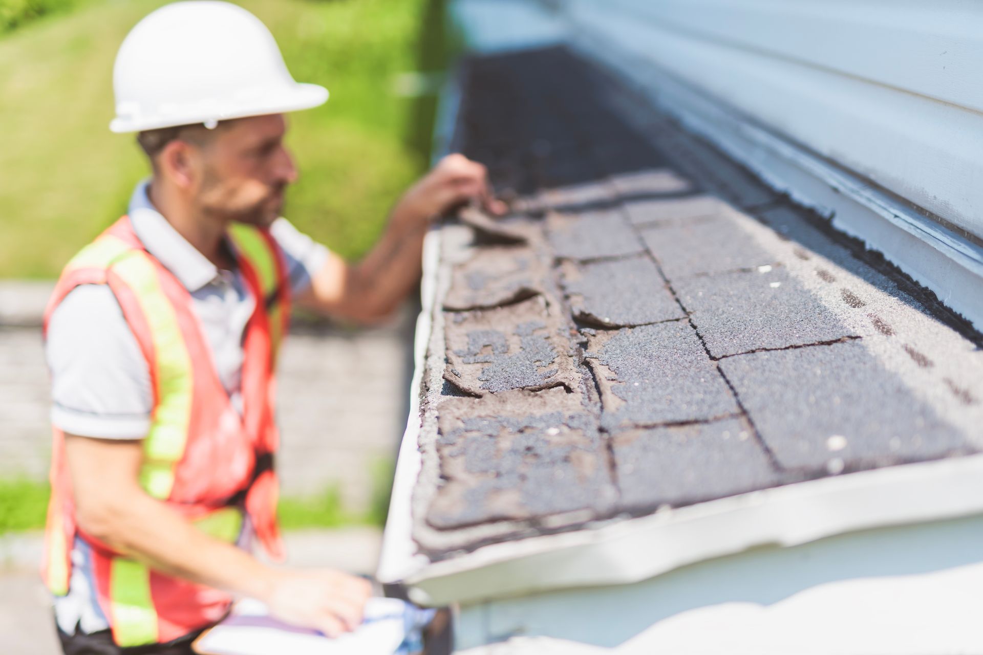 Worker in hard hat inspecting damaged roof shingles beside a house