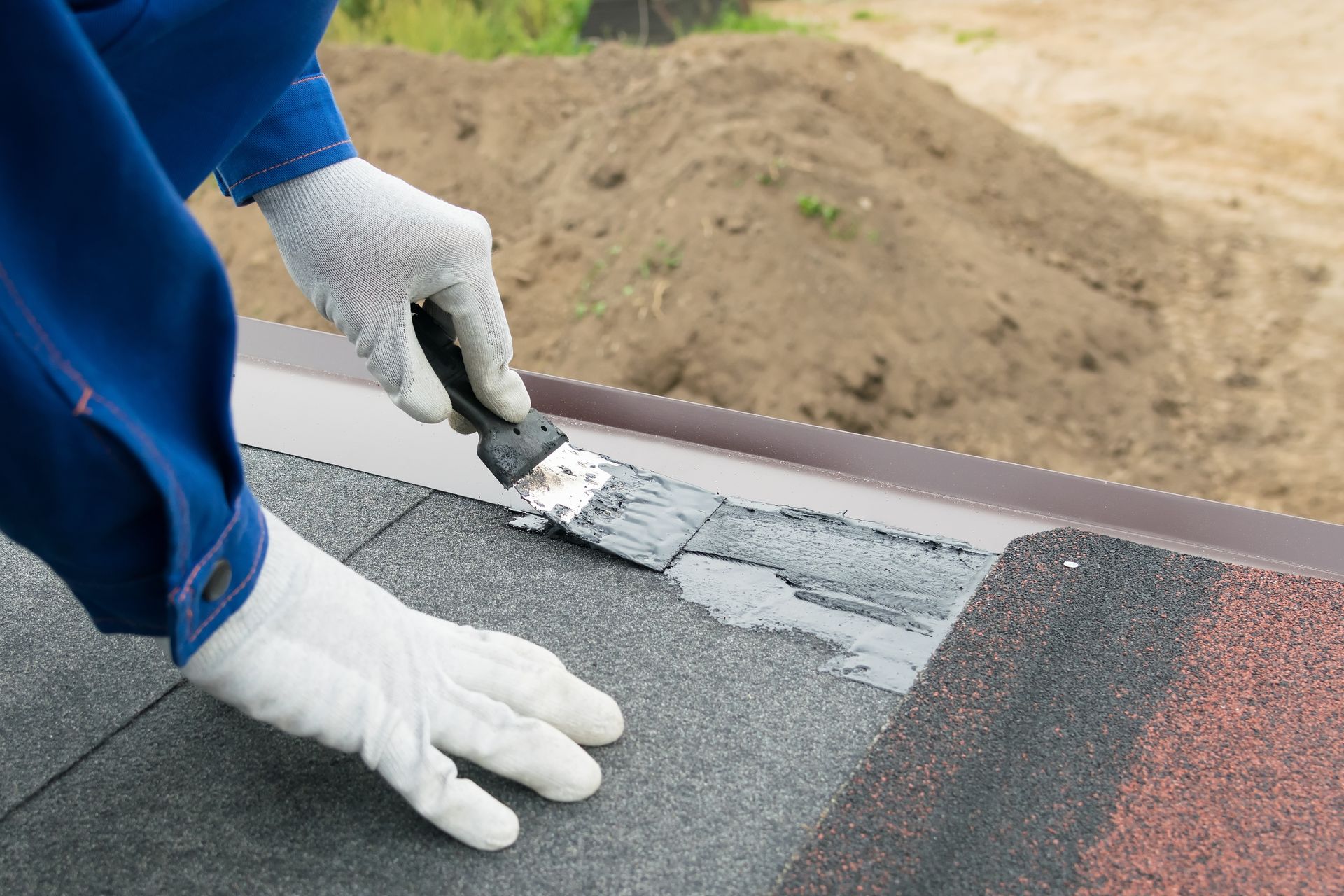 Gloved worker applying gray roofing sealant to a shingled roof seam