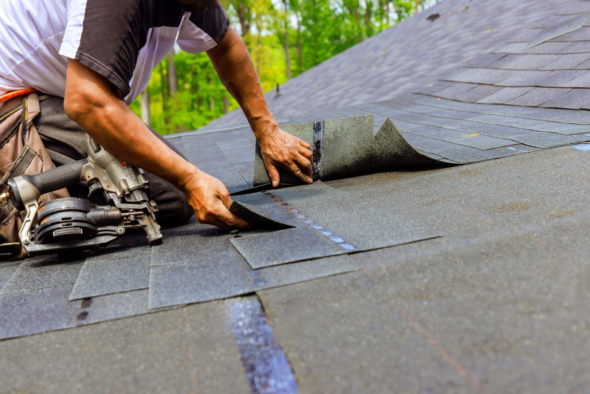 Worker installing asphalt shingles on a roof with a roofing nail gun.
