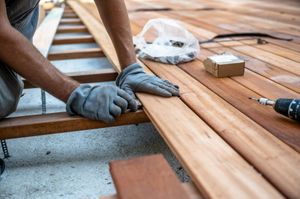 Gloved hands installing wooden deck boards with a drill and tools on a patio frame
