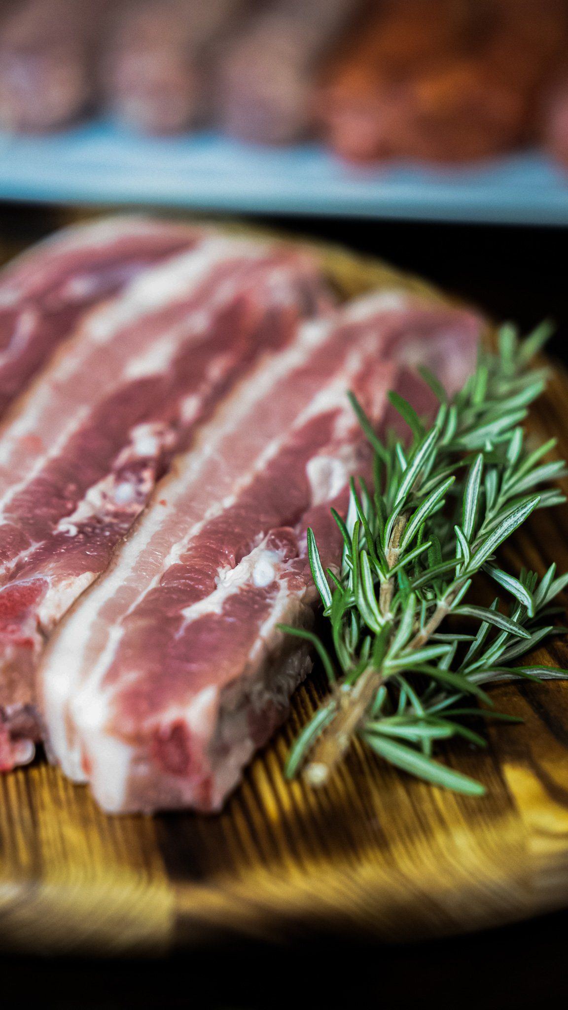 A piece of raw meat with rosemary on a wooden cutting board.