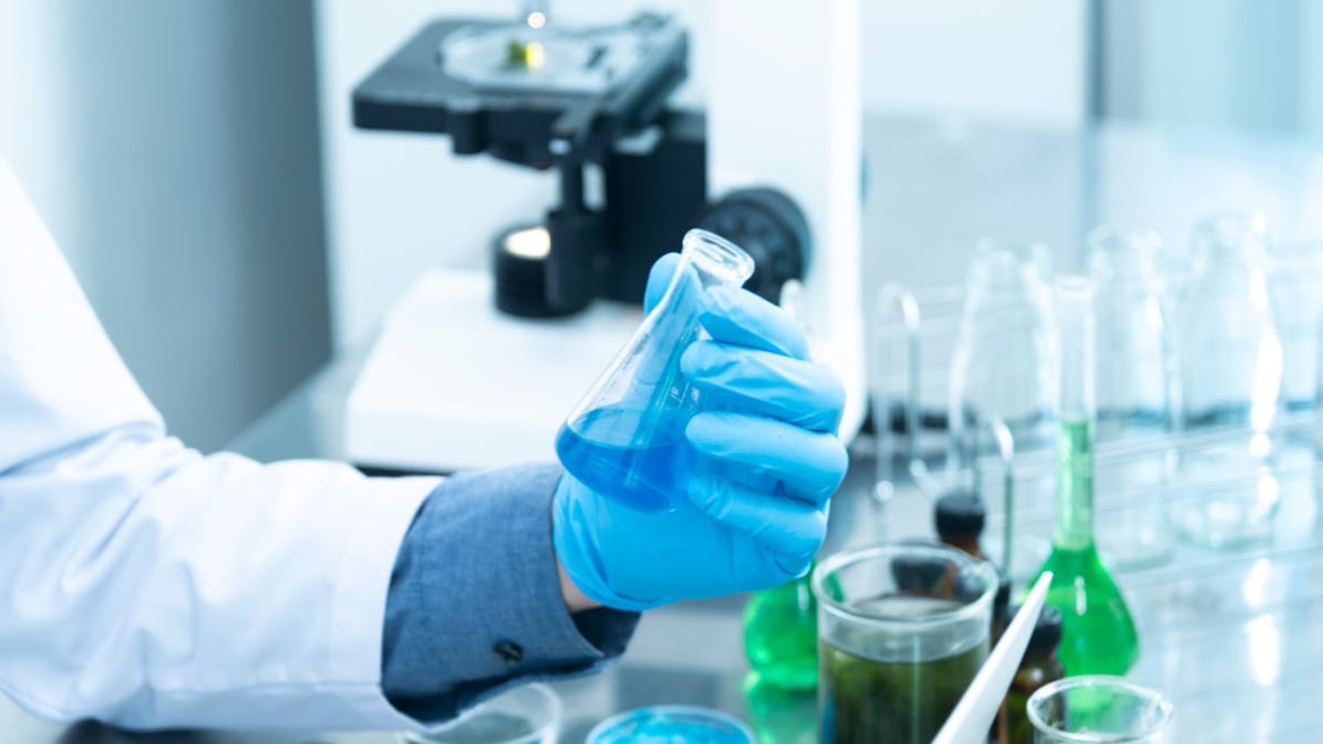 Científico con bata de laboratorio y guantes sosteniendo un líquido azul en un matraz, con un microscopio al fondo.