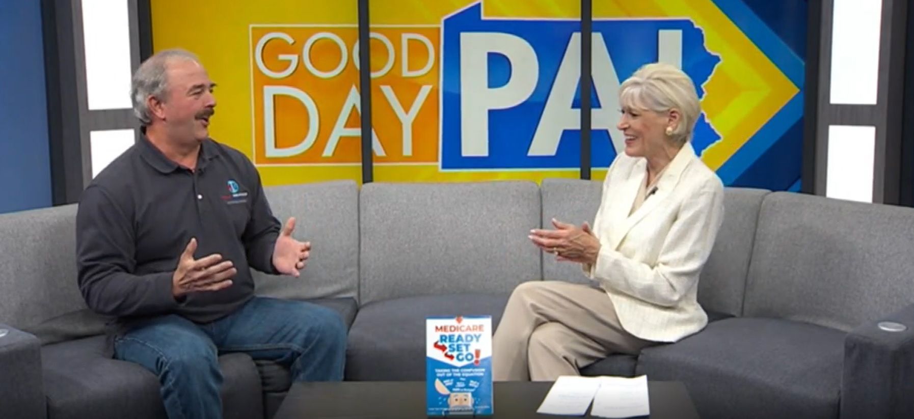 A man and a woman sitting on a couch on the set of Good Day PA, with a book placed on the coffee table in front of them.