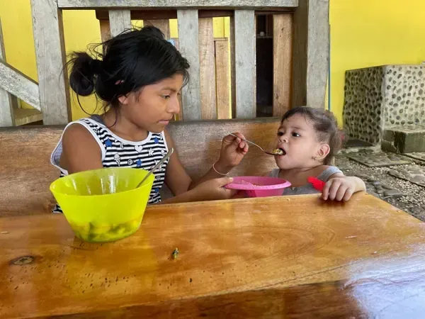 A girl feeds a toddler at a wooden table. Yellow bowl and pink plate.