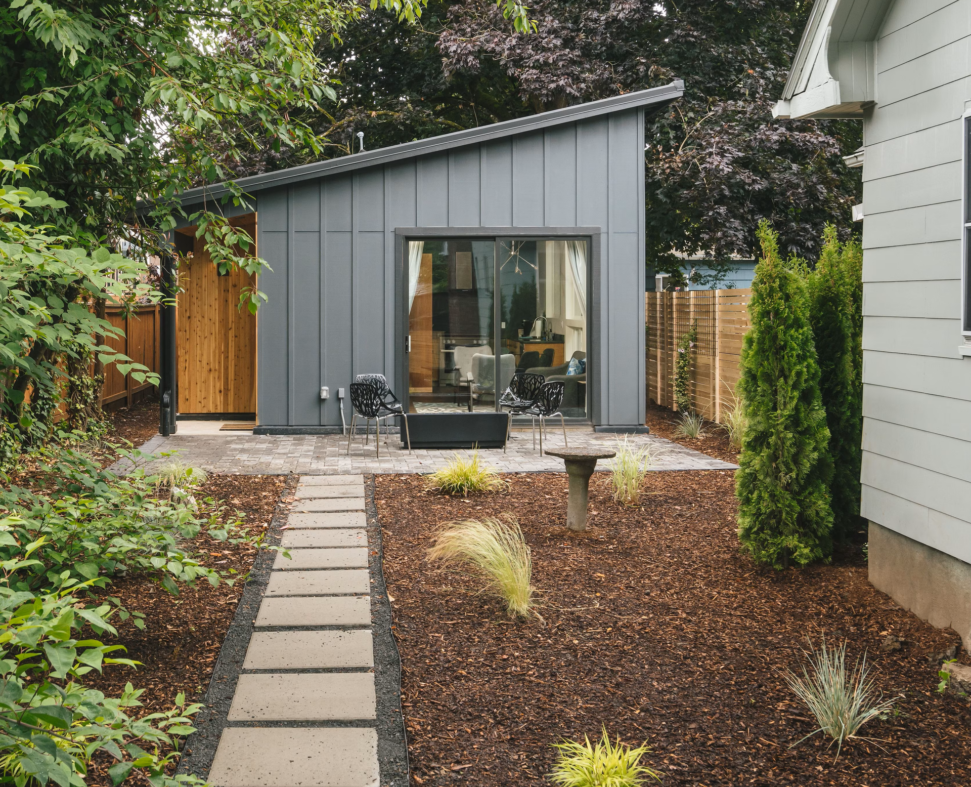 Modern gray shed with sliding glass door, patio, stepping stones, and surrounding yard.