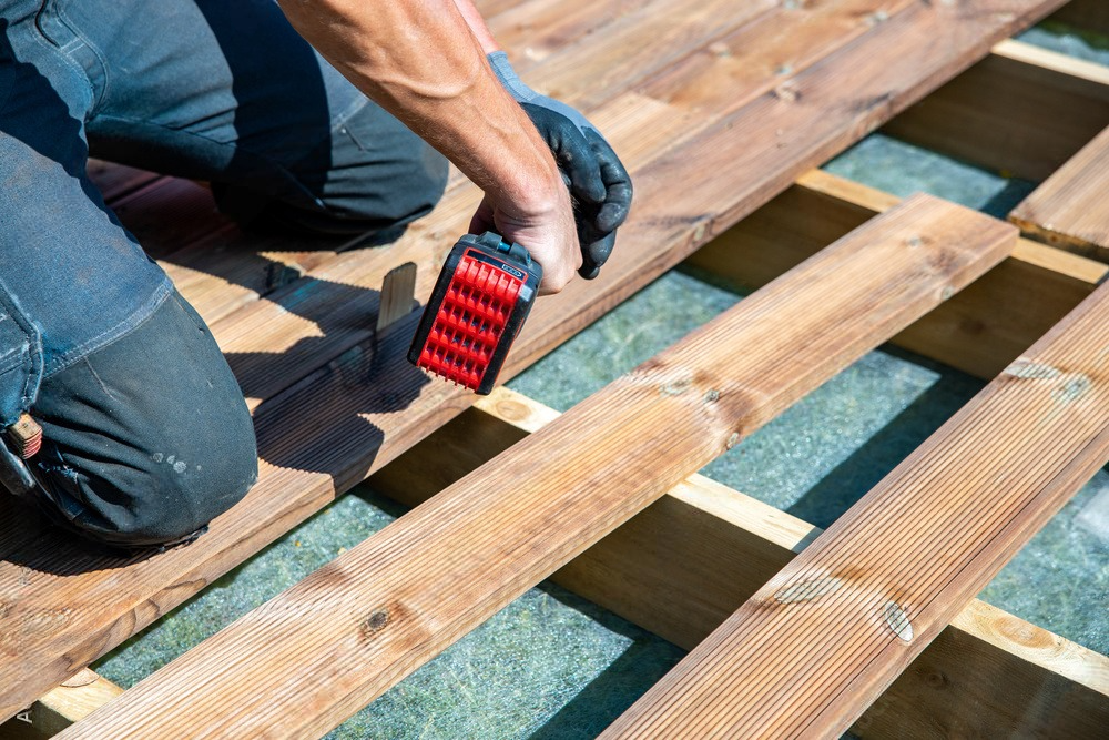 Person kneeling, installing wooden deck planks with a power tool.