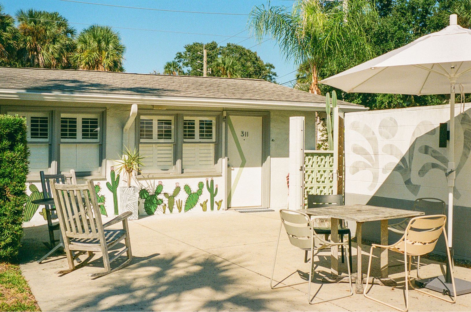 Sunlit patio with white chairs, umbrella, and a small house decorated with cactus murals.