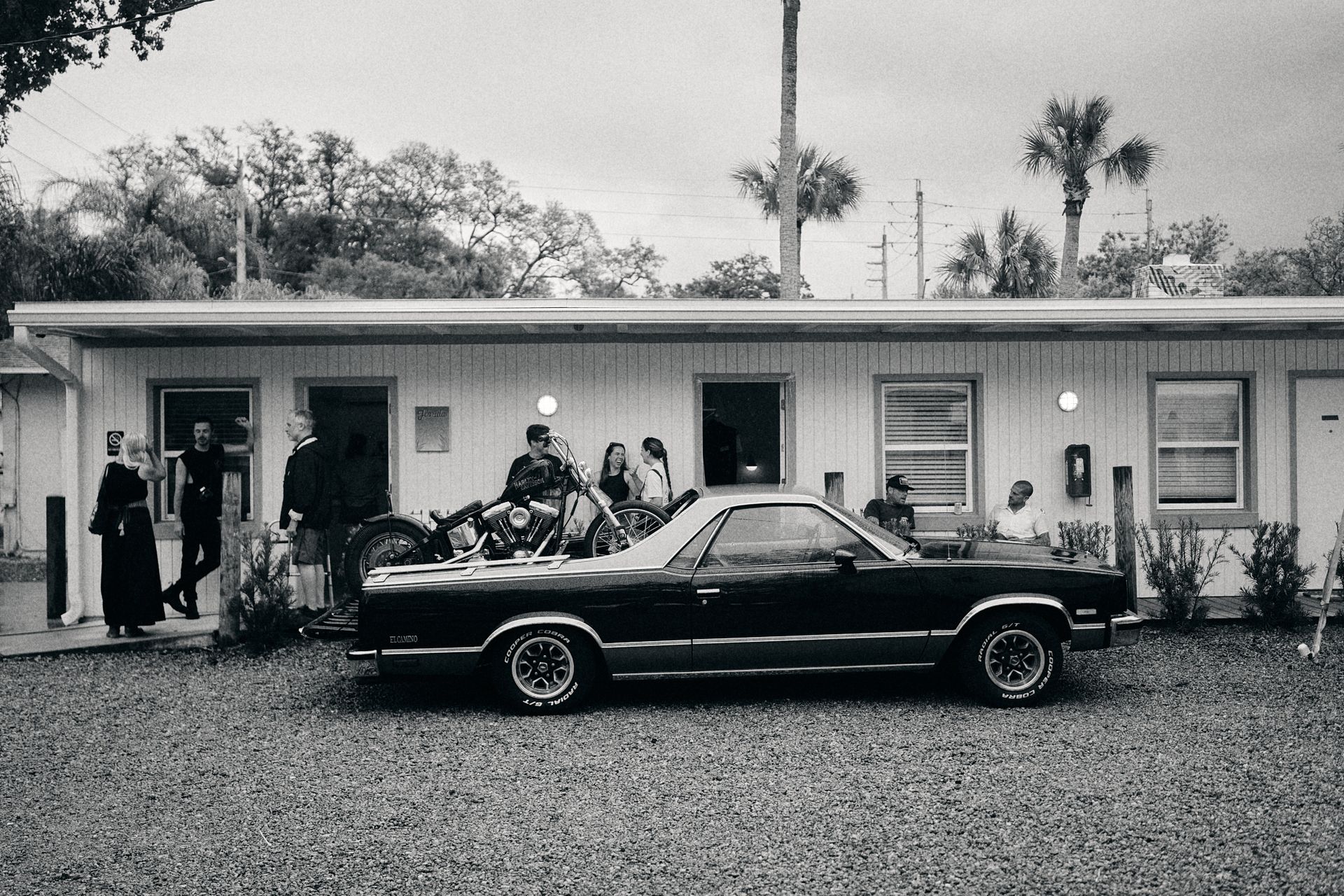Vintage black car parked in front of a motel, with people gathered outside the rooms.