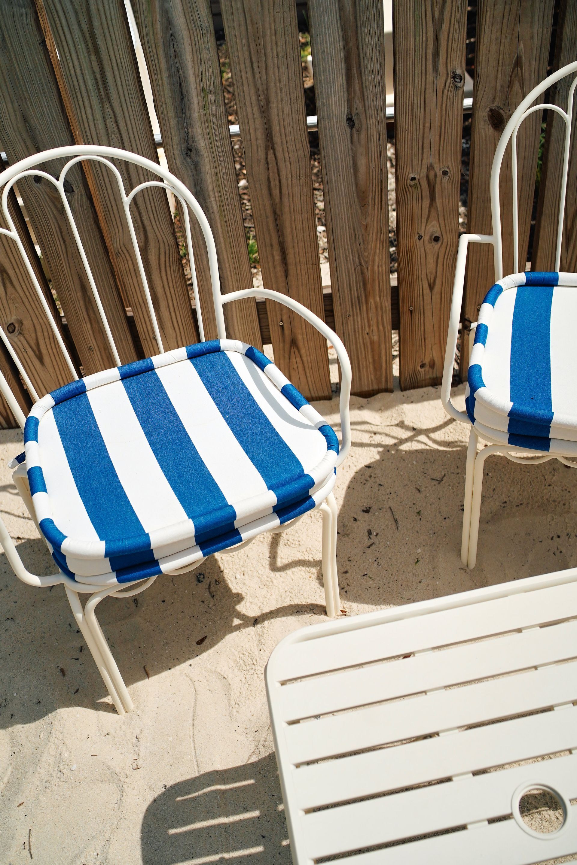 Two white metal patio chairs with blue-and-white striped cushions on a sandy deck by a wooden fence