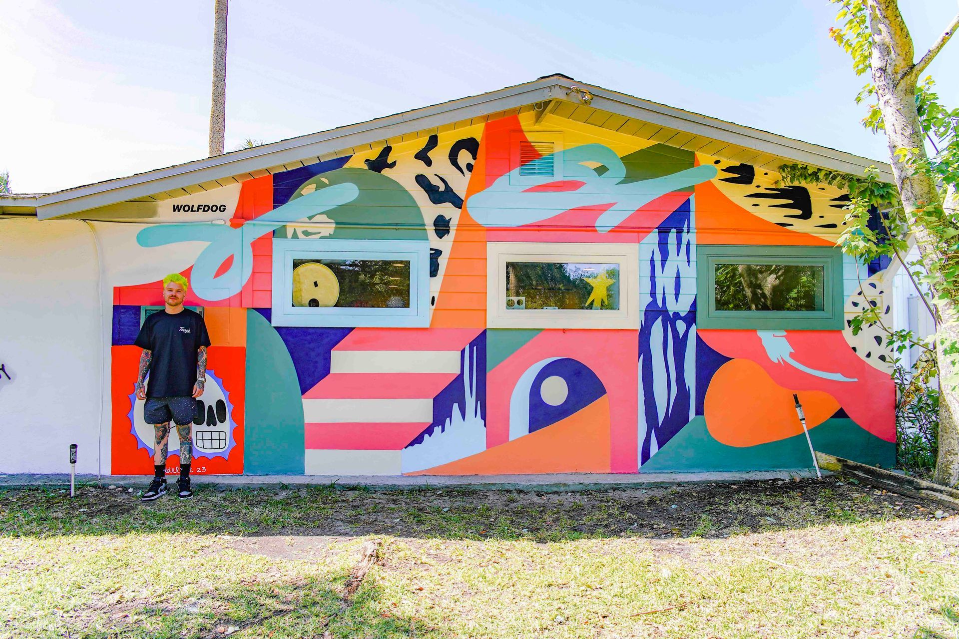 Person standing beside a brightly painted mural on a small building with windows and a slanted roof.