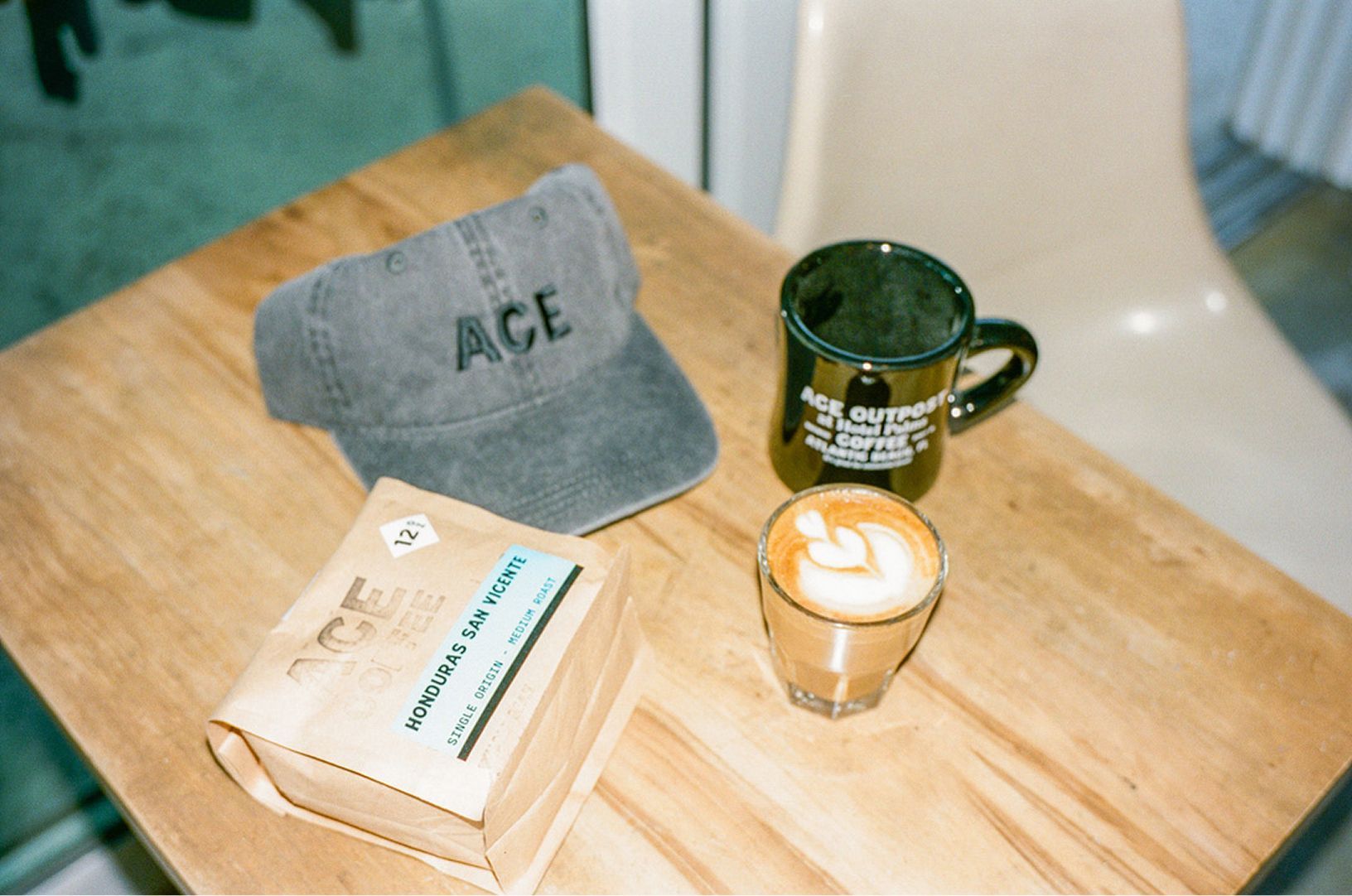 Gray ACE cap, green mug, glass of latte, and a book on a wooden table by a chair