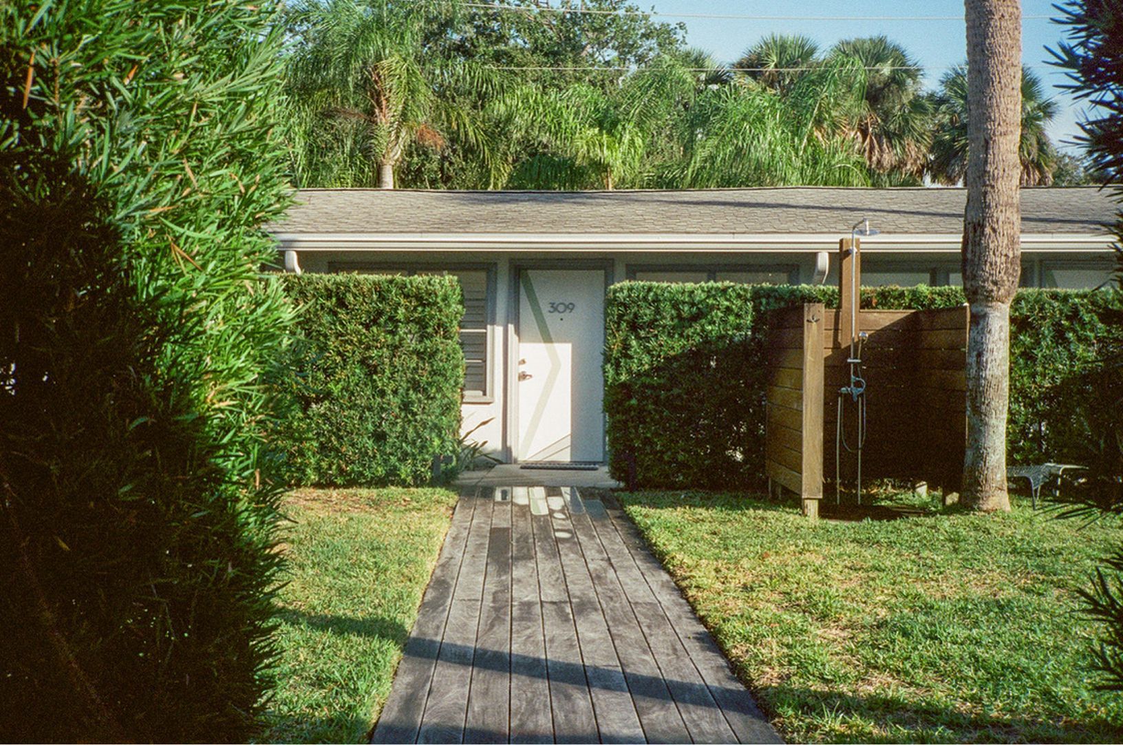 A house entrance with a white door, covered walkway, and wooden path surrounded by greenery.