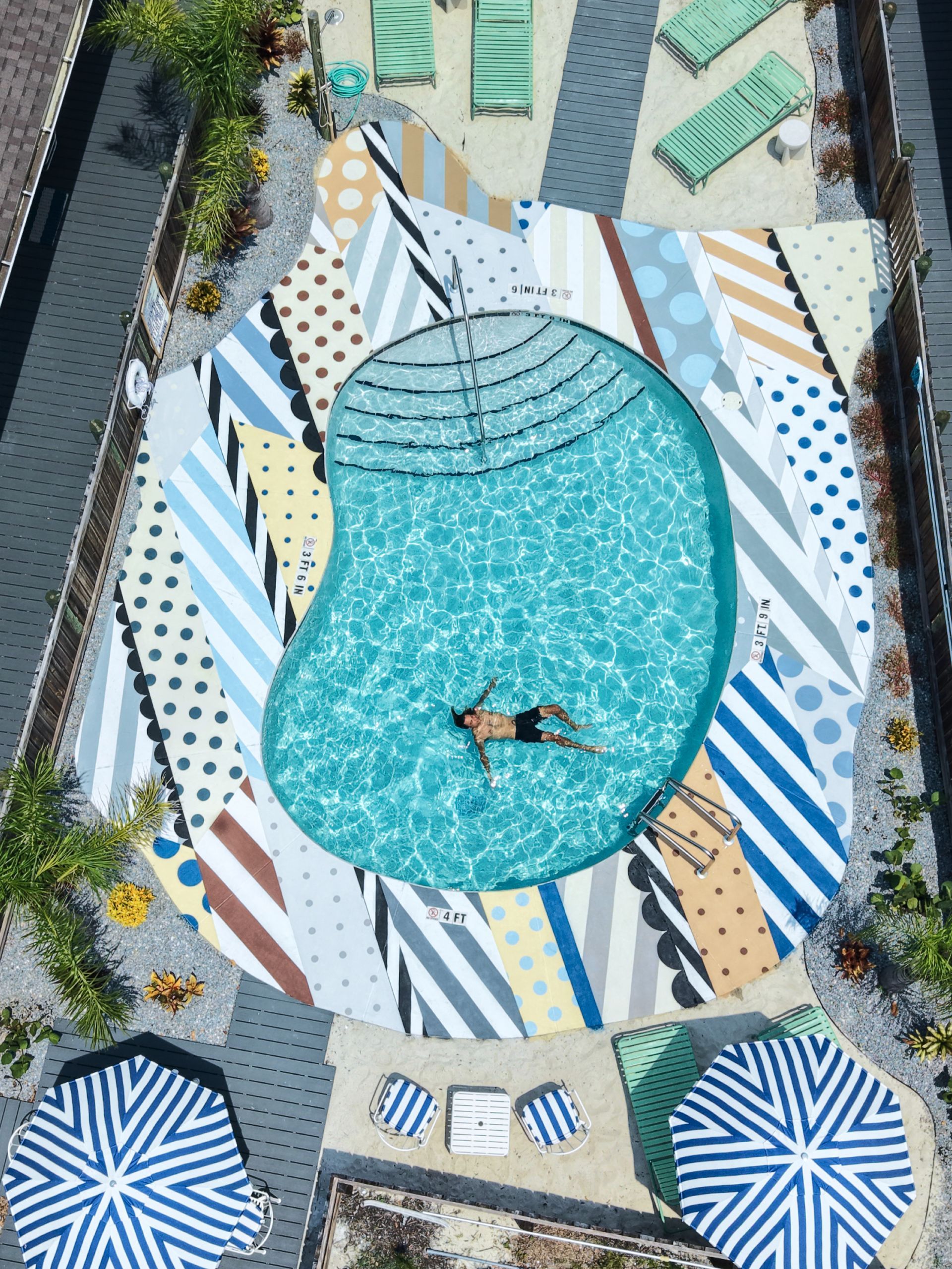 Overhead view of a turquoise pool with a swimmer, surrounded by striped loungers and umbrellas.