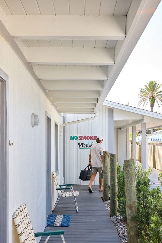 Covered outdoor walkway with a person carrying bags near a door marked “No Shoes Please.”