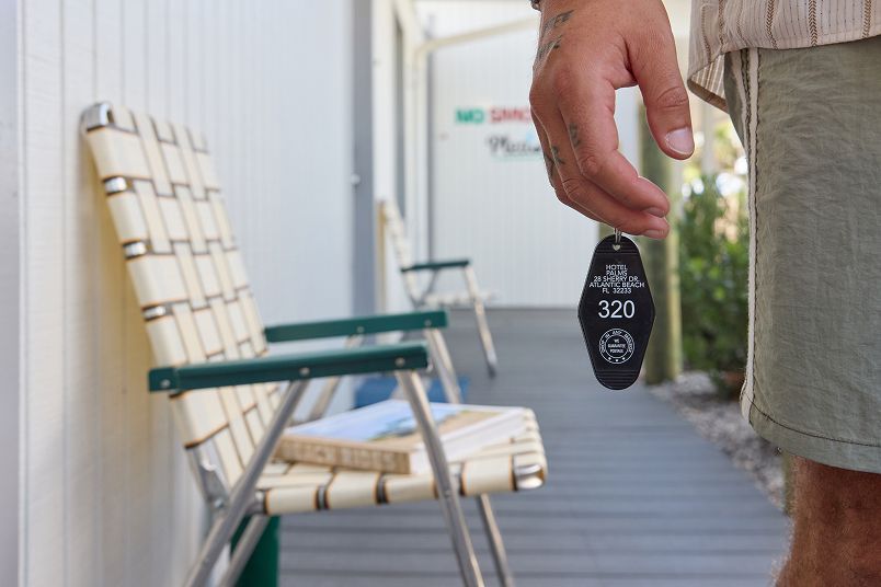 Hand holding a black hotel room key fob on a motel walkway with chairs beside a door
