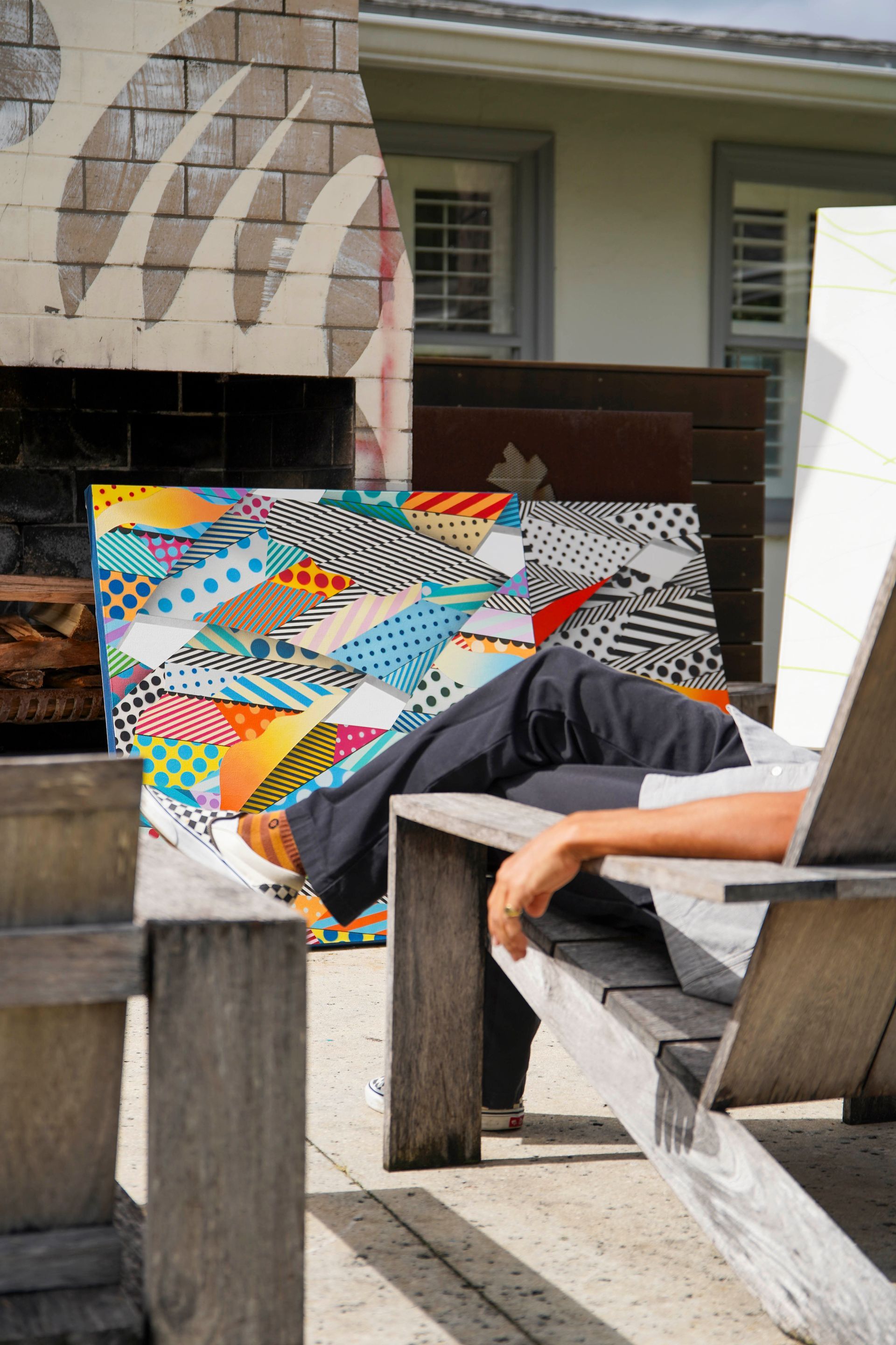 Person reclining on a wooden chair beside a colorful quilt display outdoors.