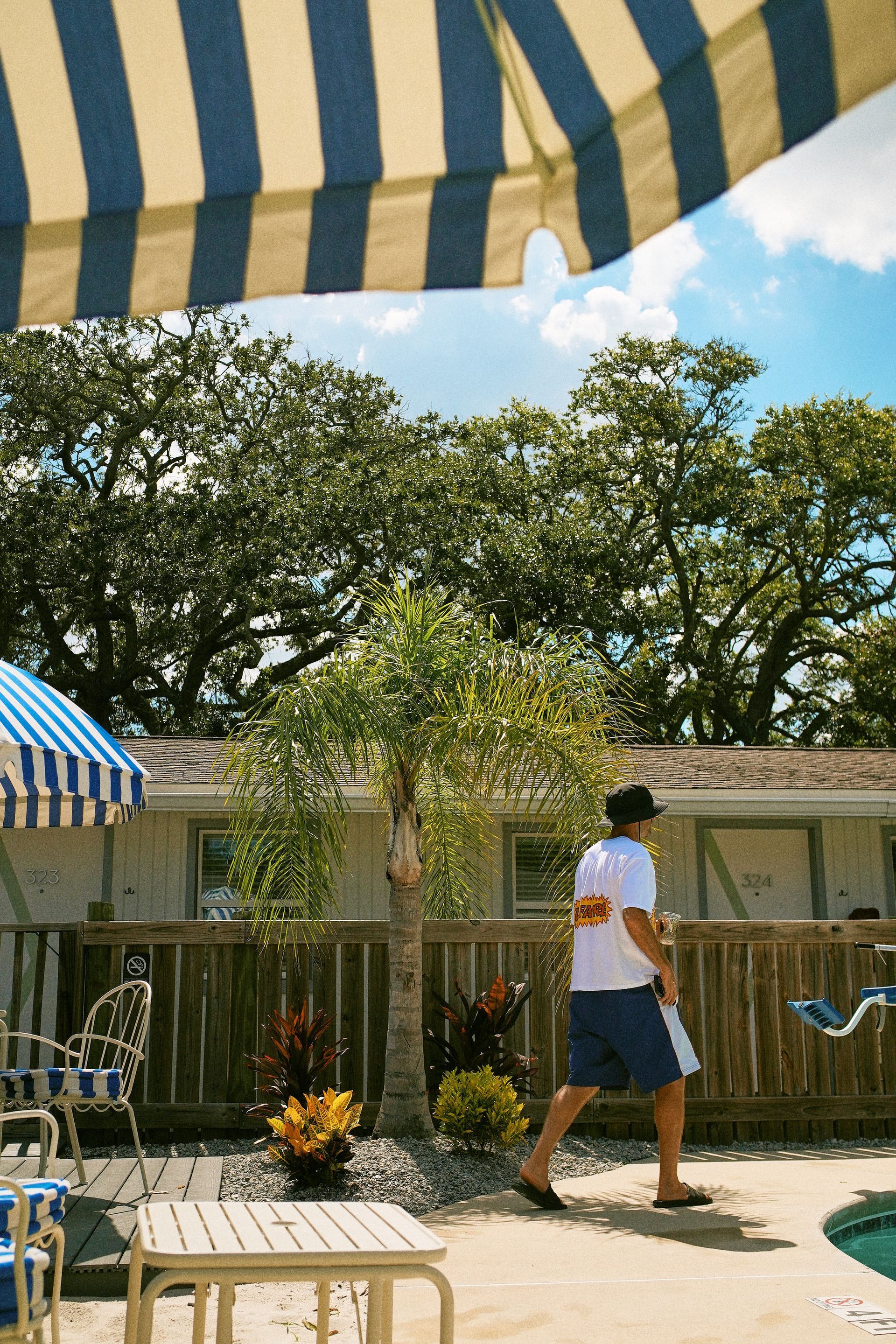 Person walking by a pool under blue-and-white striped awnings, with trees and patio chairs nearby.