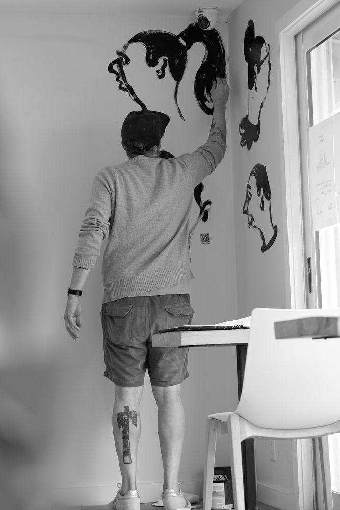 Person painting a black mural on a wall in a bright room beside a window.