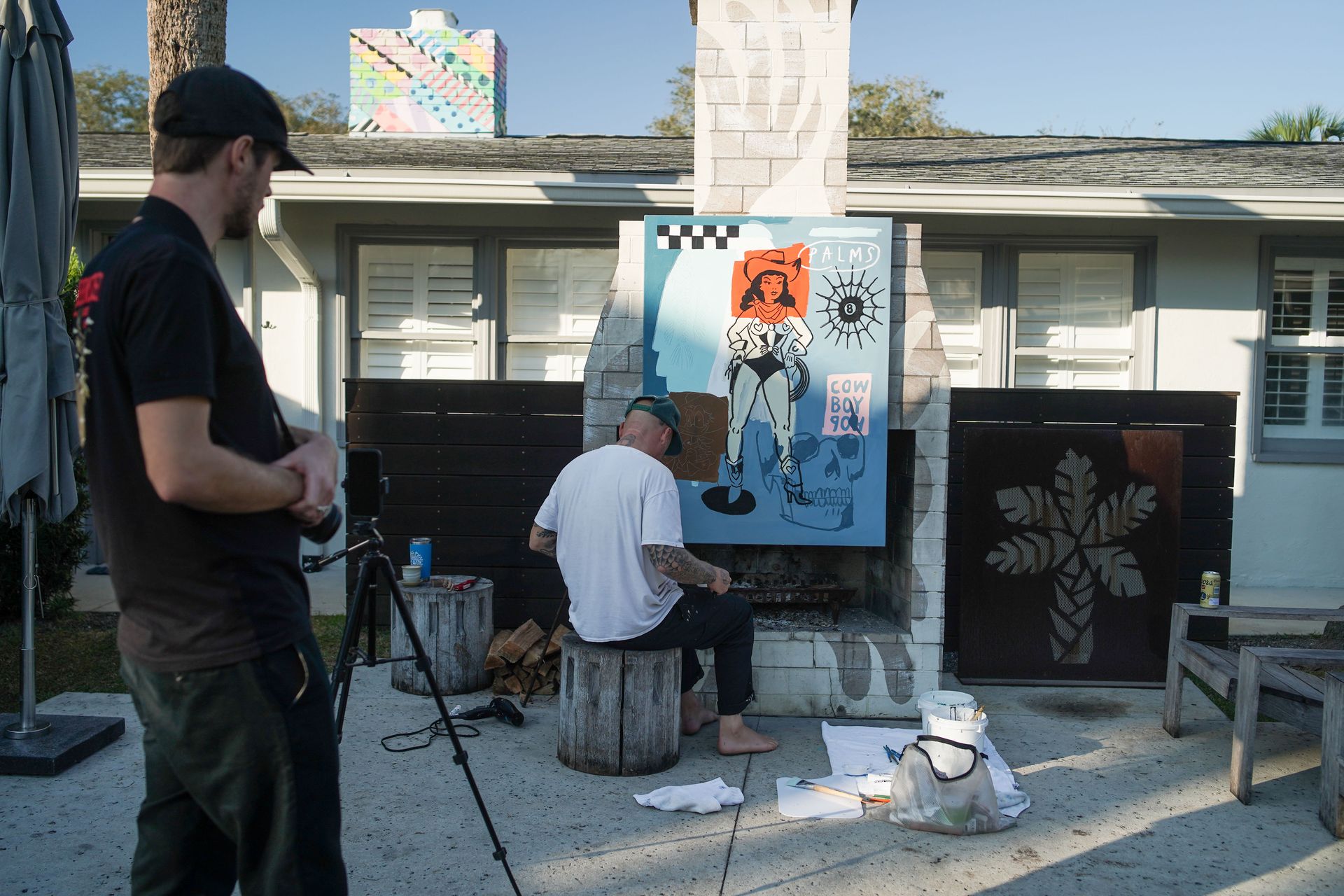Two people painting on easels in a courtyard, with canvases, paint supplies, and a house in the background