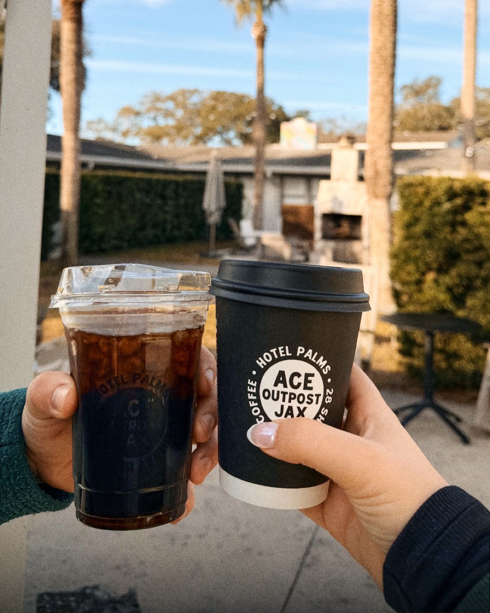 Hands holding an iced coffee and a paper cup in a sunny outdoor courtyard