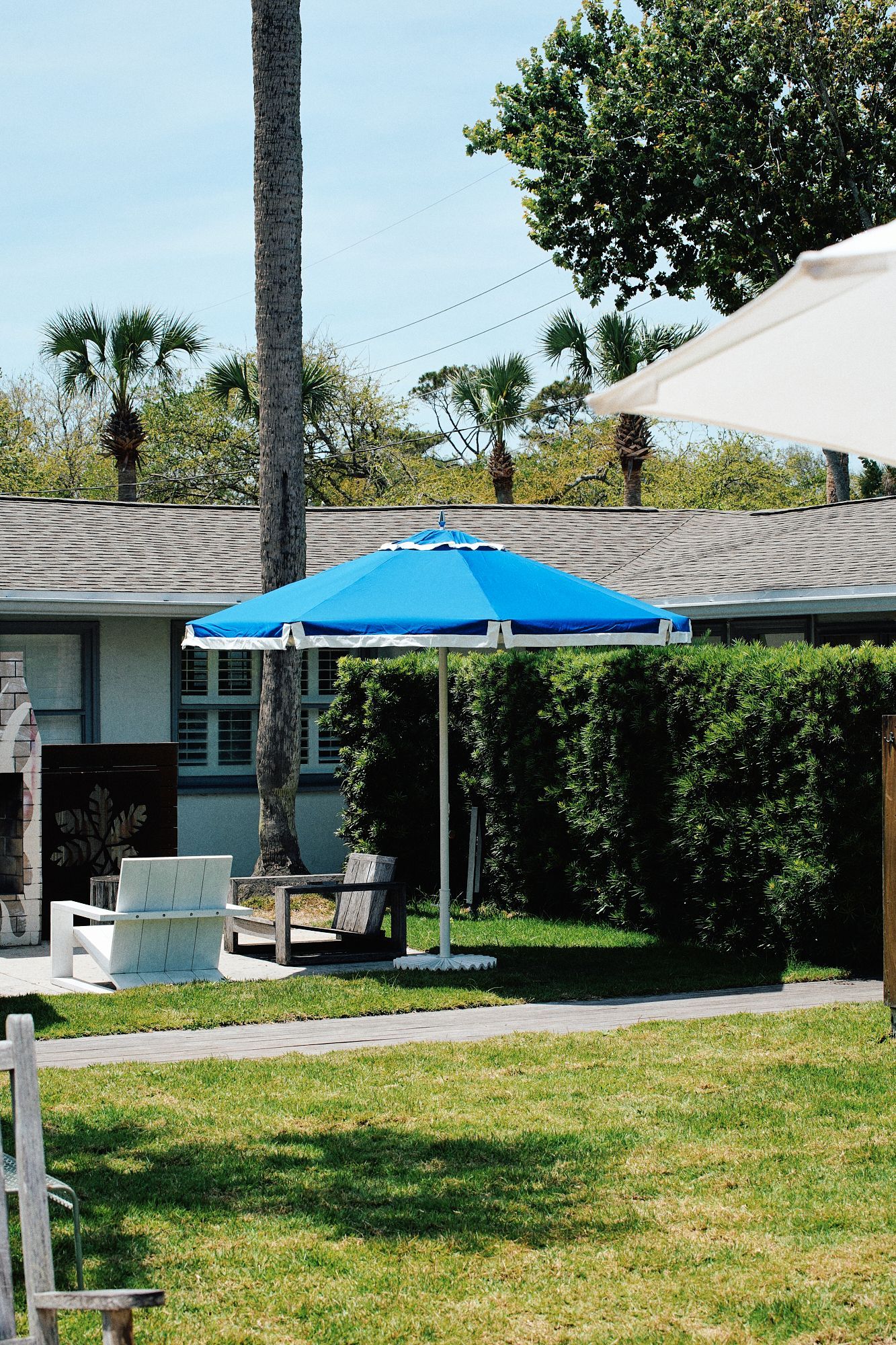 Sunny backyard patio with blue umbrella, lawn, lounge chairs, and a low house in the background