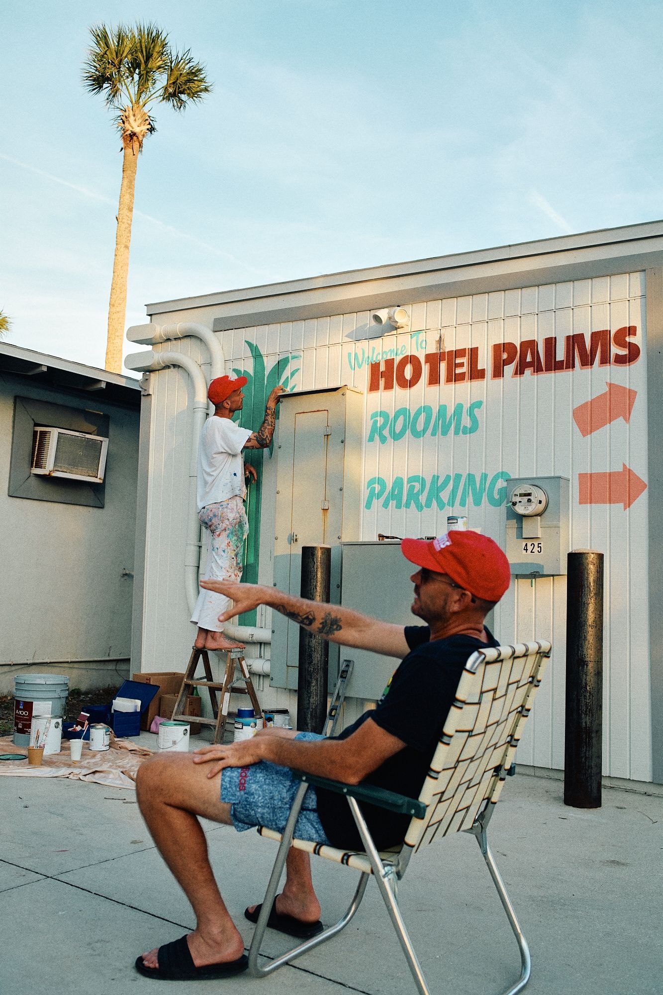 Man painting motel sign while another man sits nearby outside Motel Palms.