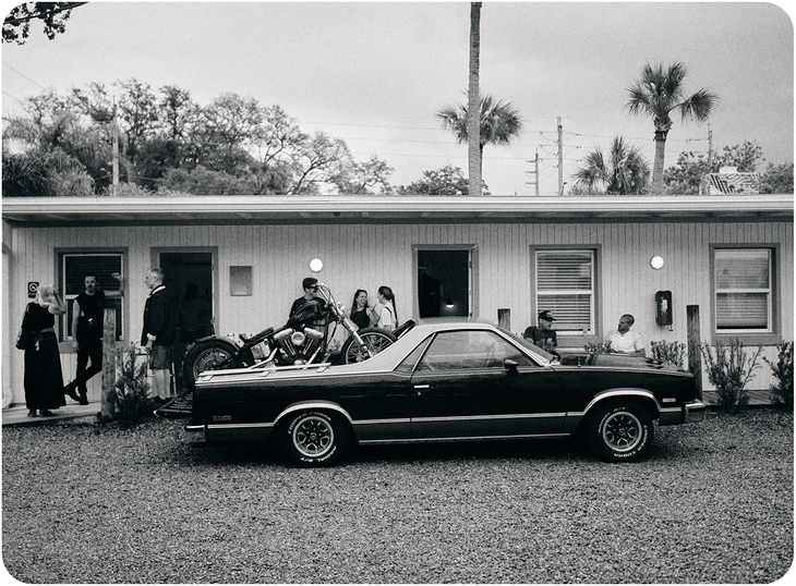 Vintage black car parked in front of a motel, with people gathered outside the rooms.