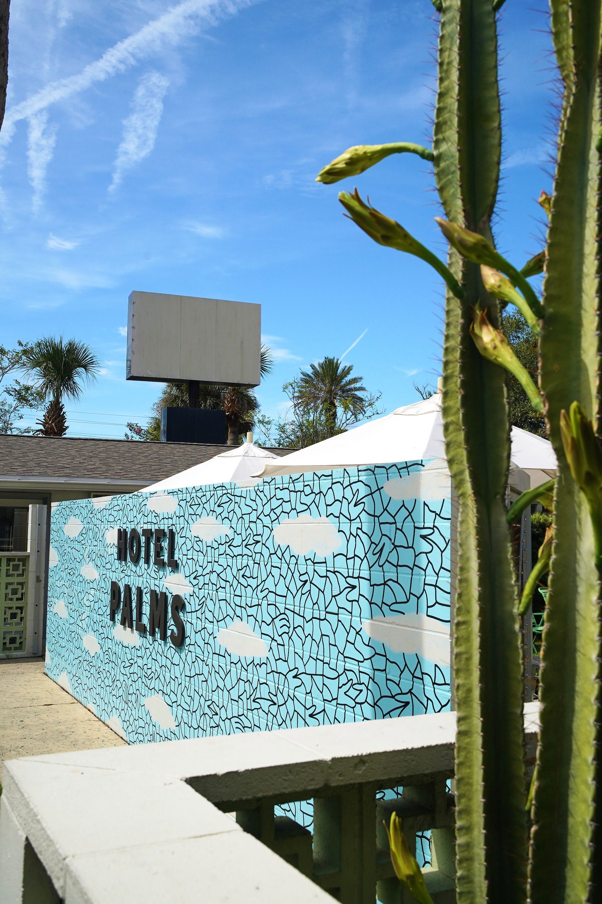 Outdoor sign with turquoise mosaic wall, white canopy, and tall cactus under a blue sky.